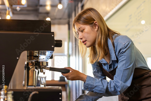 Asian woman entrepreneur making coffee from machine while standing by coworker at cafe. Female barista making coffee in coffee shop counter. Takeaway food. Professional coffee making. Small business.
