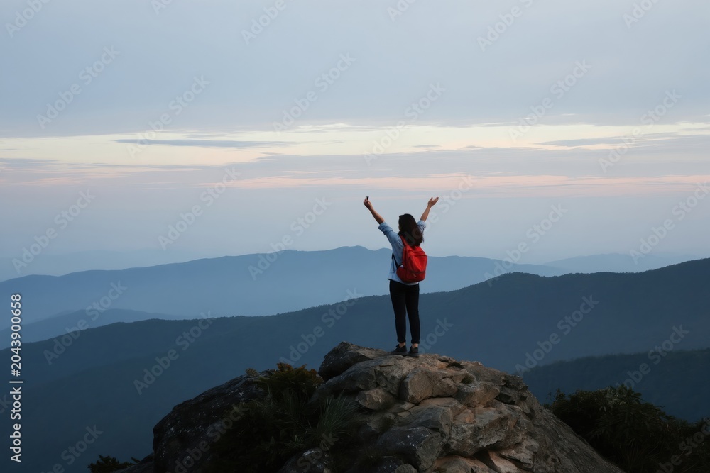 Obraz premium Woman with red backpack stands on mountain peak arms raised in victory. Hiker enjoys sunrise view over misty blue hills. Solo traveler celebrates achievement in nature.
