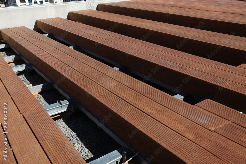 Naklejka premium New wooden deck boards laid on metal substructure. Outdoor terrace construction uses brown timber planks. Urban building background shows construction process for new exterior living space.