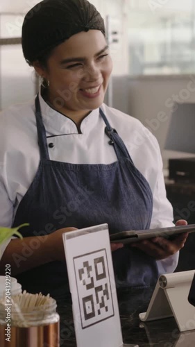 Vertical video: Answering customer greeting, barista in apron tapping tablet, smiling at counter QR
