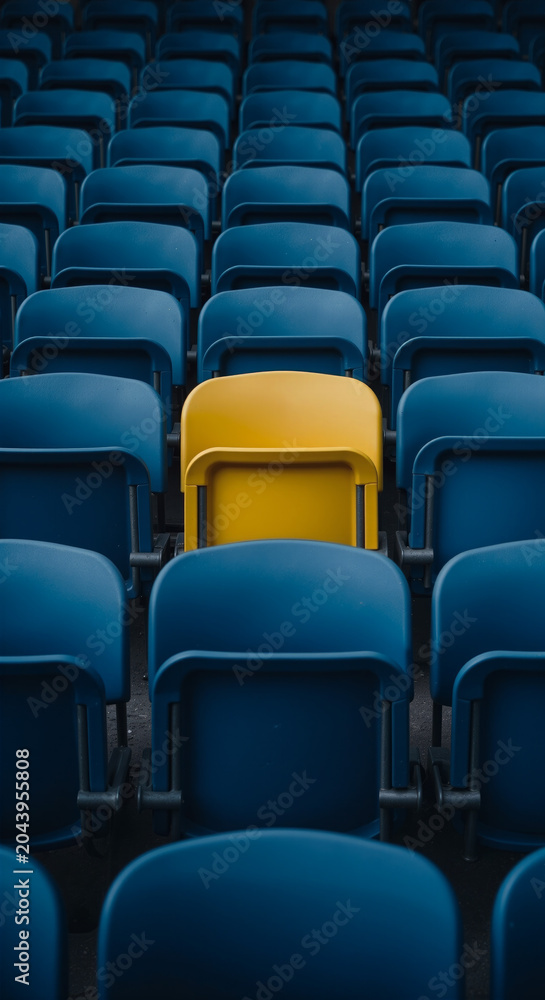 Fototapeta premium A single yellow stadium seat stands out amongst rows of blue seats in a sports arena.