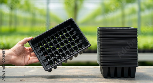 Hand holding a black plastic seedling tray next to a stack of identical trays in a greenhouse nursery setting