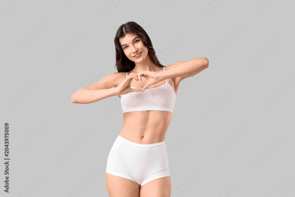 Fototapeta premium Pretty young woman in underwear making heart with her hands on light background