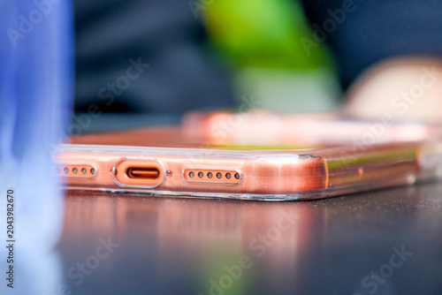 Glossy phone rests on dark surface. Metallic edges gleam under soft light. USB port and speaker grilles visible. Blurred background suggests casual setting. Ideal for tech, lifestyle, or product ads