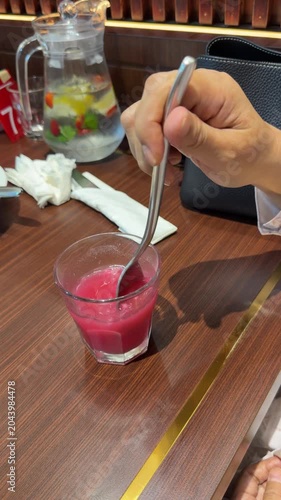 Hand Stirring Pink Drink in Glass on Wooden Restaurant Table