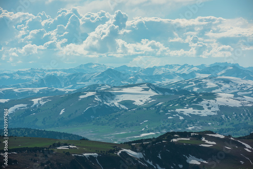 Aerial sunlit top view above forest hills and big snowy mountain range under lush clouds in changeable weather. Scenic hilly alpine landscape with sunlight and shadows. Partly cloudy in high mountains