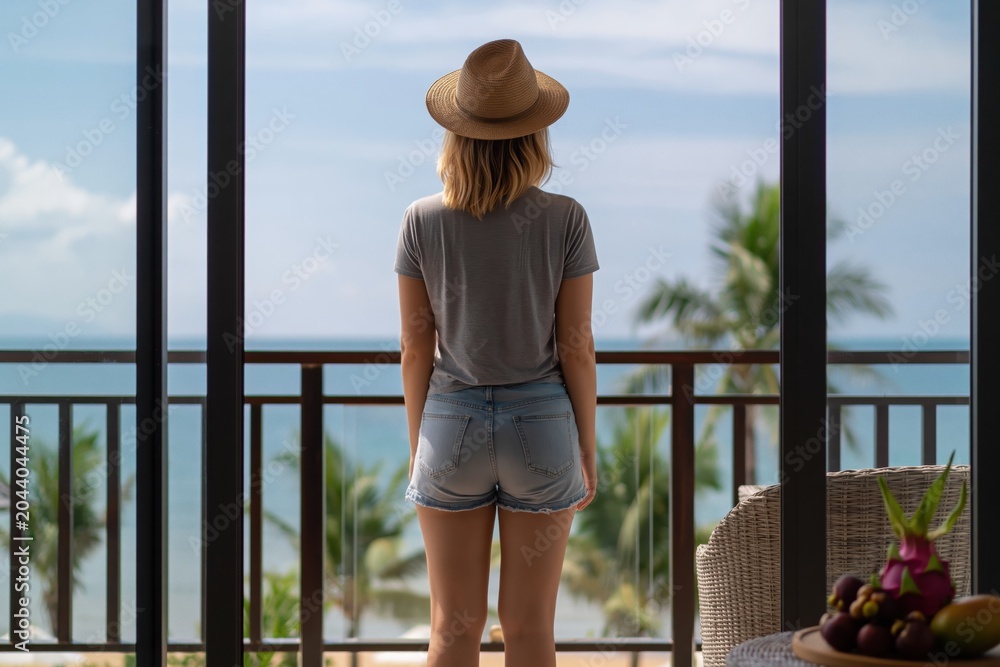 Fototapeta premium Woman standing on hotel balcony rear view of tropical ocean and palm trees female person