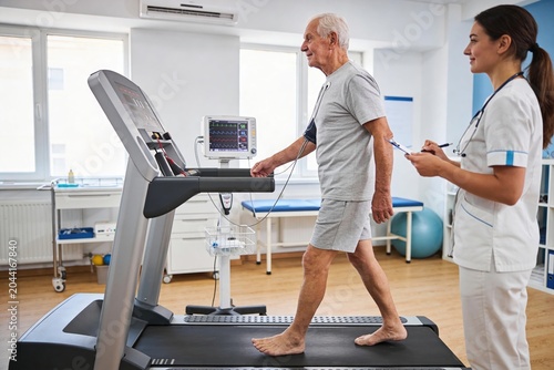 Senior man performing cardiac stress test on treadmill with nurse monitoring vitals in a bright medical rehabilitation clinic