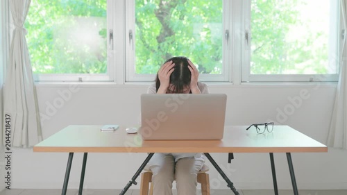 Asian woman holding her head looking distressed while working on laptop