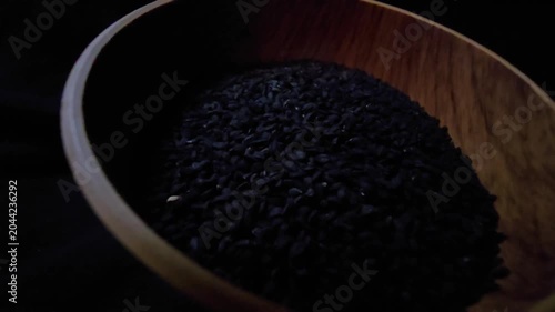 Macro cinematic shot capturing black nigella seeds (Nigella sativa), also known as black caraway, spilling out from a rustic wooden bowl onto a wooden table. The sharp focus and shallow depth of field