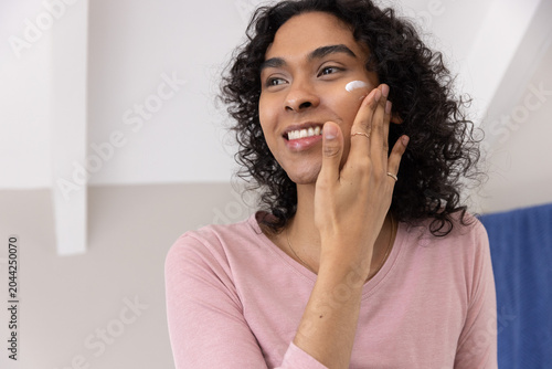 Non-binary person applying white cream to cheek at vanity, wearing pink top, rings and necklace