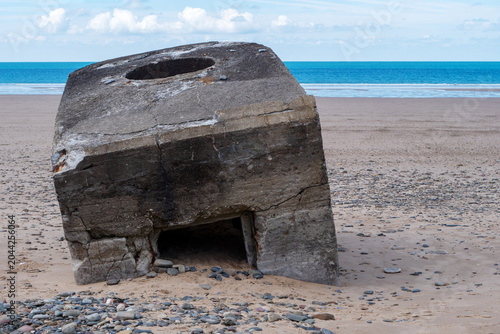 Weathered concrete bunker sits on sandy beach. Blue ocean stretches behind structure. Normandy, France