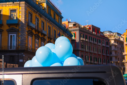 Lively city street in Naples, Italy with massive cluster of blue balloons emerging from behind a vehicle. Vibrant historic architecture creates a cheerful, festive urban atmosphere