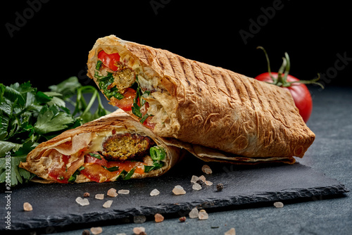 Gourmet vegan shawarma wrap cut in half with plant-based meat and fresh vegetables on black slate board, side view of delicious meatless burrito or doner kebab on dark background