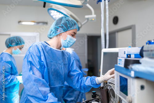 Foto Profile of focused female anaesthesiologist in blue sterile gown, mask, and cap, using gloved hand to adjust medical monitor in operating room, blurred surgical team background