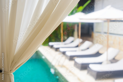 Private cozy resort swimming pool with lounge chairs and umbrellas behind white curtain in focus. Luxury summer vacation background.