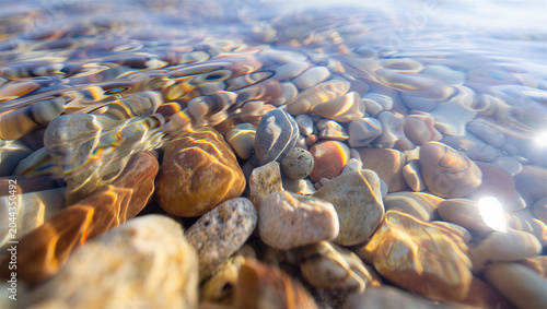 A close up view of colorful smooth river stones submerged under clear, shallow water with light reflecting and refracting on the rippled surface