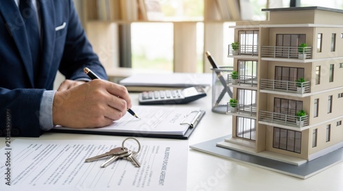 Businessperson signing paperwork with pen on clipboard beside metal keys and a detailed apartment building model on desk; calculator nearby, sunlight through window