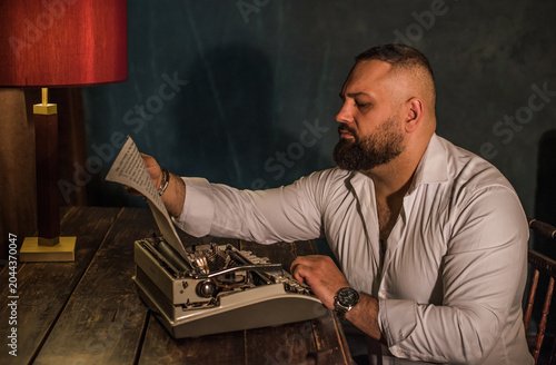 Serious bearded man writer sitting at a vintage desk and working with a retro typewriter, male author looking at a manuscript in a dark atmospheric room, classic noir style storytelling concept