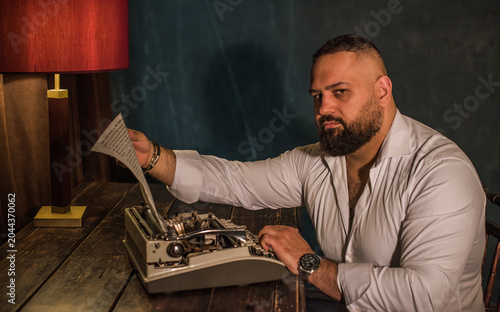 Serious bearded man writer sitting at a vintage desk and working with a retro typewriter, male author looking at a manuscript in a dark atmospheric room, classic noir style storytelling concept