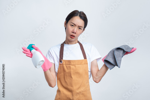 Canvas Print Asian woman in an apron and pink rubber gloves, looking confused and holding cleaning tools, isolated on white background