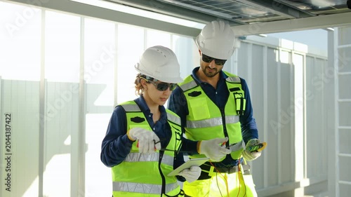 Two professional solar energy engineers in safety gear inspect panel installation using a digital multimeter and tablet at a renewable energy site, discussing technical green maintenance work.