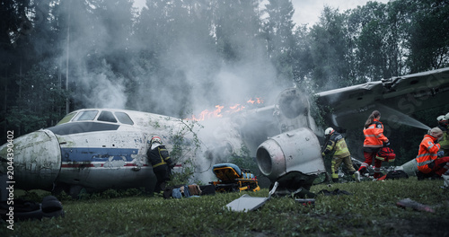 Foto Paramedics and Firefighters Work Amid Thick Smoke at a Forest Airplane Crash, Accessing Broken Fuselage Sections, Suppressing Flames, Coordinating Urgent Rescue Around Scattered Wreckage and Debris