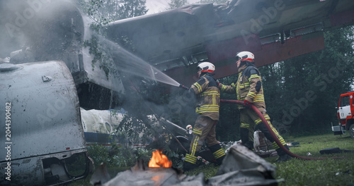 Photos Firefighters Direct Hose Streams Onto a Shattered Airplane Fuselage in a Wooded Crash Zone, Cooling Metal Wreckage, Suppressing Flames, and Clearing Smoke During Urgent Airplane Rescue Operations