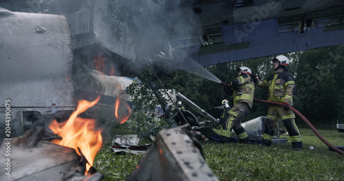 Foto Firefighters Work Around Burning Airplane Wreckage in a Forest Crash Site, Moving Through Smoke and Debris to Suppress Flames, Secure the Fuselage and Support Survivor Rescue Operations Near Trees