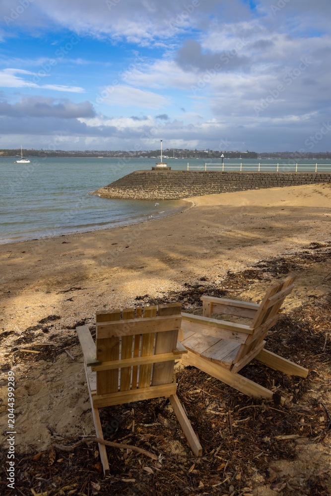 Obraz premium Two empty wooden chairs sit on the beach in Devonport, Auckland, New Zealand, providing a peaceful place to enjoy the harbor and calm sea.