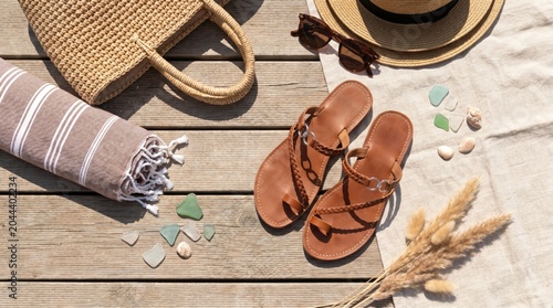 A flat lay of summer essentials on a wooden surface. Brown sandals, a woven bag, sunglasses, a straw hat, and decorative stones are arranged neatly.