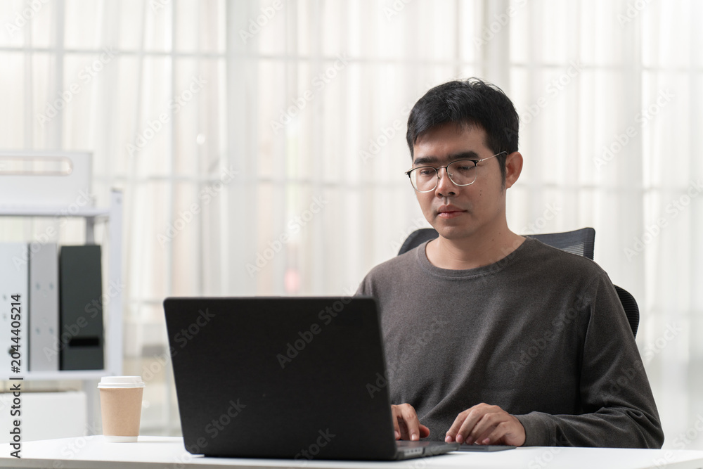 Fototapeta premium Young male professional working on laptop at office desk with beige curtain background