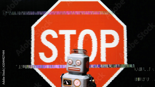 Retro robot toy stands before glitching red stop sign with digital distortion effects on black . Pixelated corruption and colorful scan lines create cyberpunk aesthetic. AI warning .
