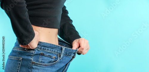 Confident woman shows loose trouser celebrating healthy weight journey success after fitness diet progress standing on blue background