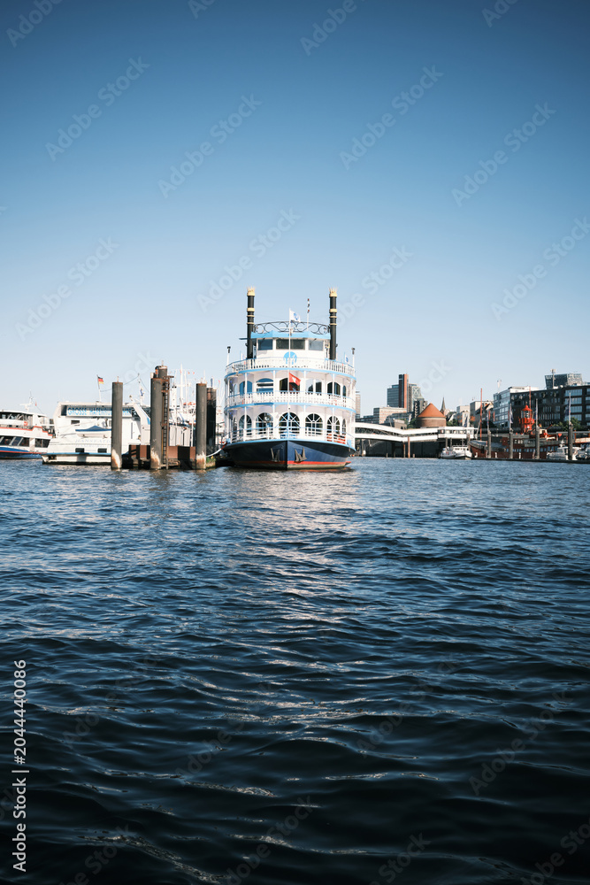 Obraz premium A large paddle steamer is docked beside a busy waterfront with other boats, harbor pylons, and modern city buildings under a clear blue sky, with rippling