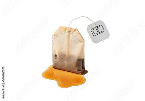 A wet, used tea bag with a paper tag resting in a small puddle of brown tea liquid, isolated against a solid black background