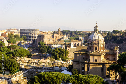 A panoramic view of the Roman Forum and the Colosseum in Rome, Italy, under a clear sky.