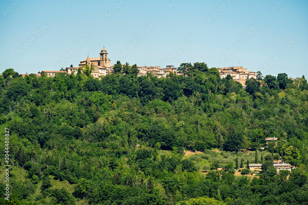 Fototapeta premium Mondaino village on its hill in Montefeltro, Italy, with the church of San Michele Arcangelo rising above green forest under clear sky