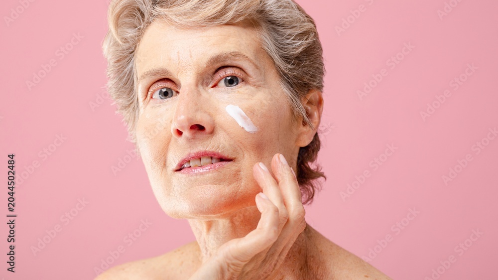 Fototapeta premium Senior white woman applying skincare cream on her face against pink background