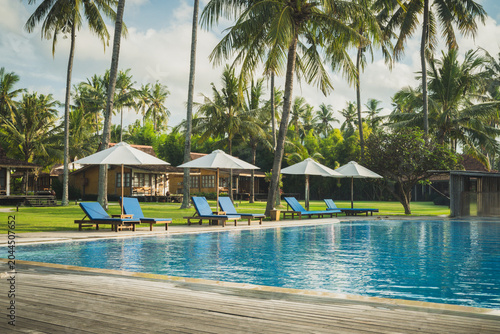 A tranquil tropical resort pool lined with blue sun loungers and white umbrellas, surrounded by swaying palm trees and lush greenery, evoking relaxation, vacation and warm sunny leisure