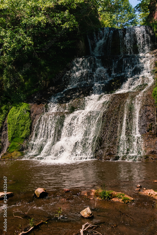Fototapeta premium A tranquil Ukrainian waterfall descending over rugged stone terraces, bordered by dense trees and reflecting the beauty of untouched nature.