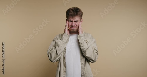 Young man experiences discomfort while holding his head and showing signs of stress in a simple room setting