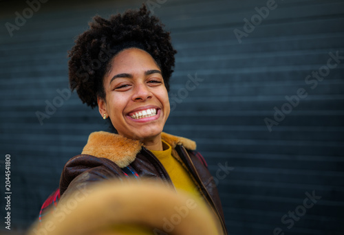 Joyful smiling young woman with afro hair wearing a stylish jacket, capturing a selfie moment outdoors against a dark urban background, expressing positivity and happiness