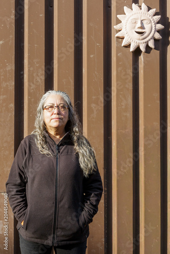 Older woman with serious expression with hands in pockets, looking at the camera, illuminated by sun, sheet metal wall and a decorative sun hanging in background, dressed casually in black clothing
