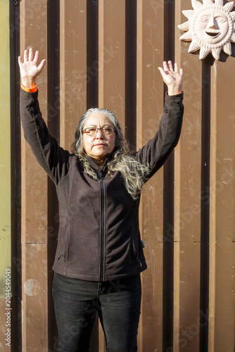 Adult woman with long gray hair standing with her arms raised, metal wall with a decorative sun hanging in the background, dressed casually in black clothing, taking a break while playing
