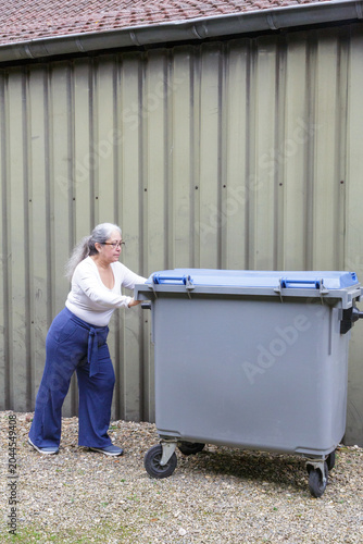 Female worker collects and pushes a wheeled mobile container of industrial waste on a farm, rough gravel terrain, integrated waste management or industrial waste collection services