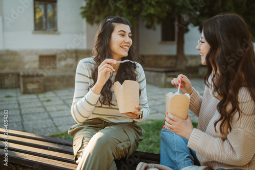 Young women friends laughing and eating takeaway outdoors