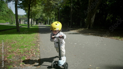 Canvas-taulu Toddler riding scooter on park path wearing yellow helmet, slows down and stops
