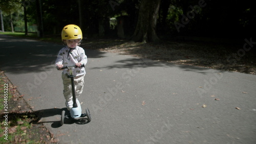 Quadro em tela Toddler riding scooter on park path wearing yellow helmet, slows down and stops