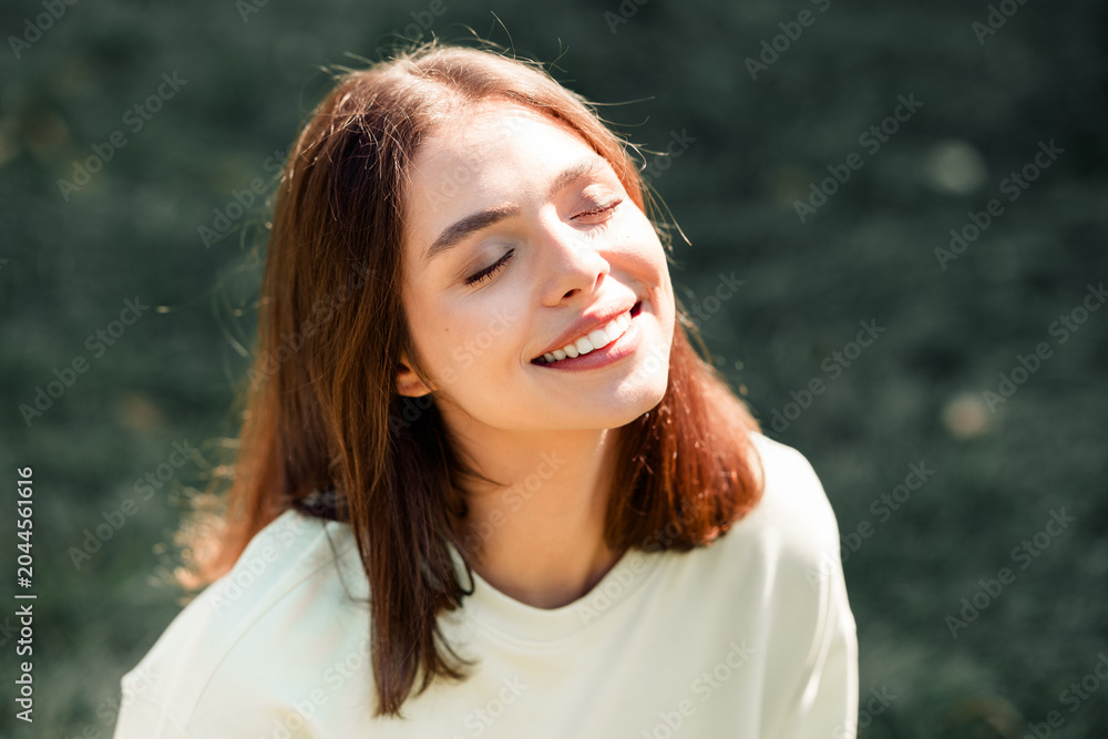 Naklejka premium Young woman smiling with closed eyes enjoying sunlight outdoors in a relaxed casual portrait